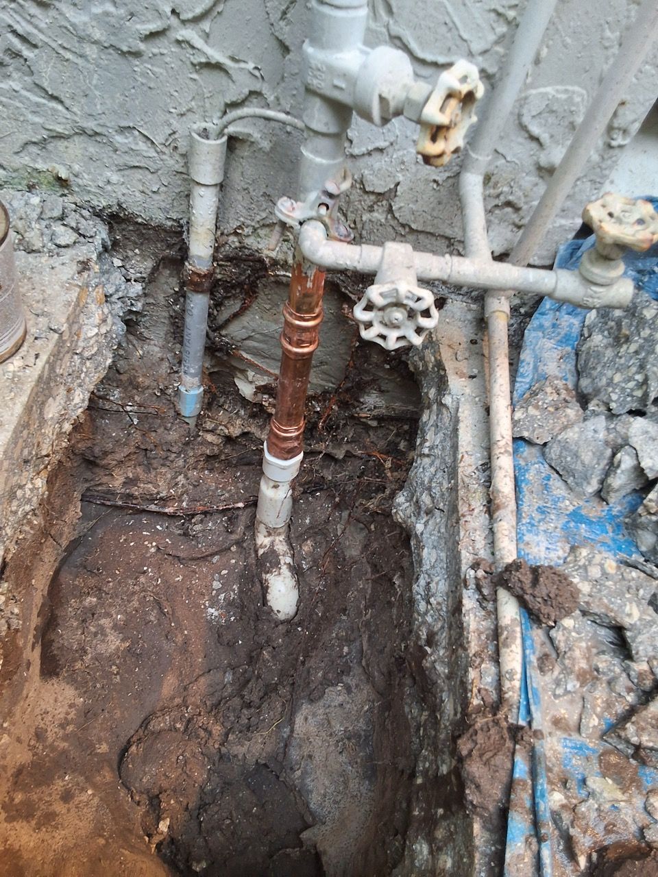 Exposed underground pipes and valves in a muddy trench beside a stucco wall