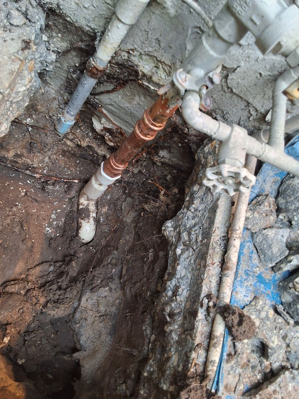 Damaged underground pipes exposed in a muddy excavation trench beside a wall