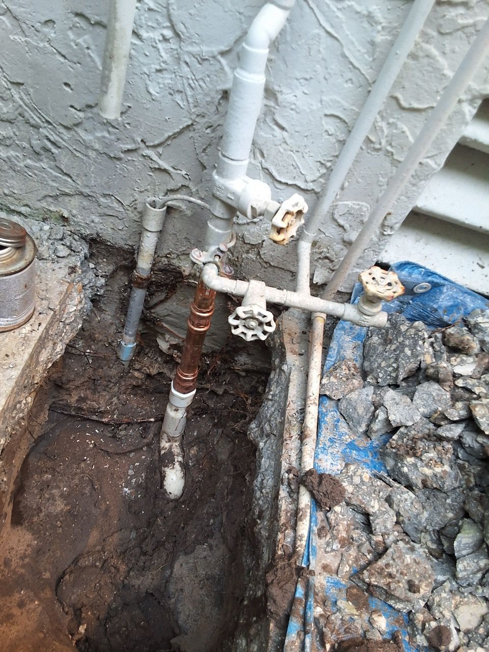 Plumbing pipes and a small excavated hole beside a house wall, with exposed soil and debris.