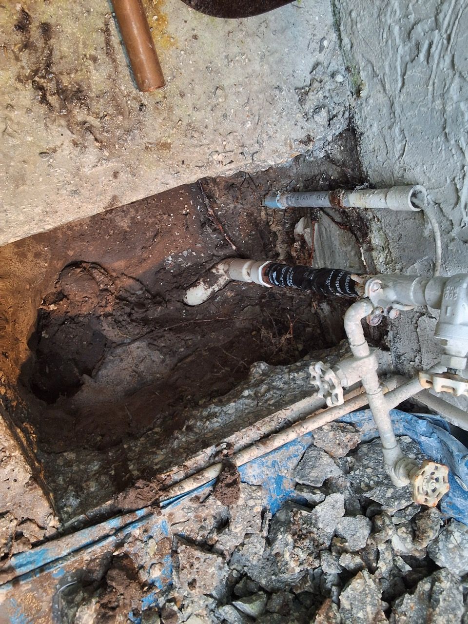 Excavation site with exposed pipes, a dark soil trench, and a white pipe spraying water near broken ground.