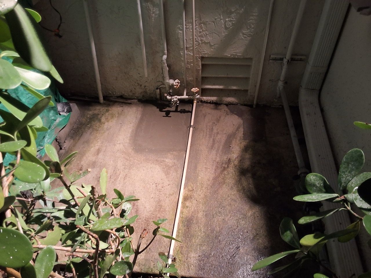 Concrete utility pit with pipes and a valve against a metal wall, lit by sunlight from above.