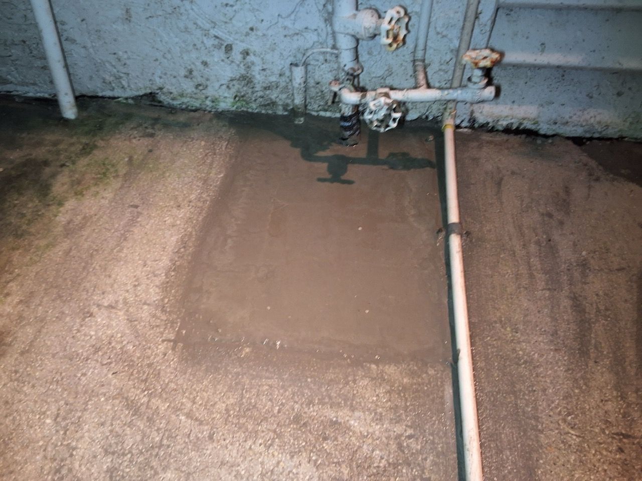 Two metal pipes and valves on a wall above a wet, muddy concrete floor