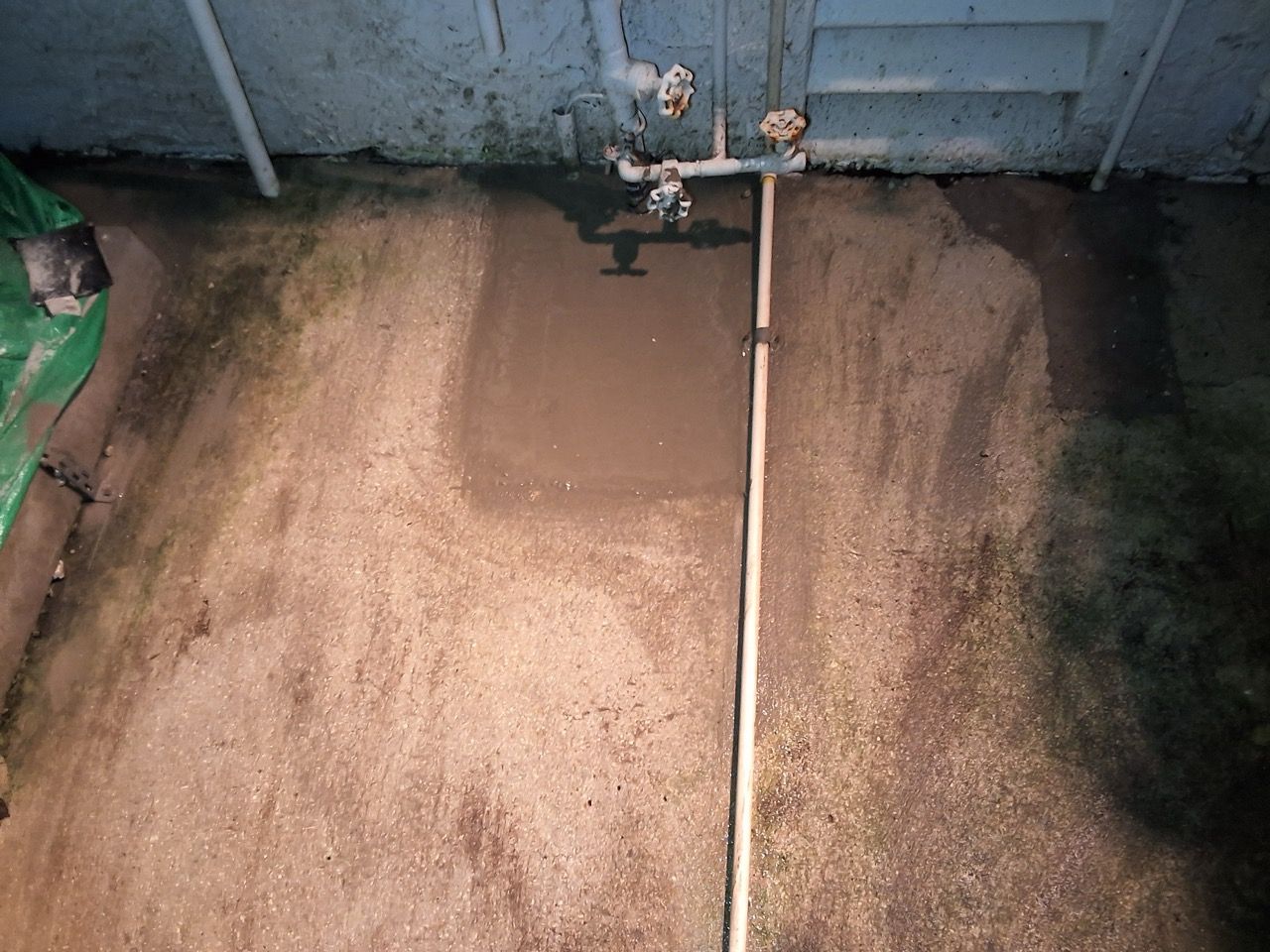 Dirty concrete floor by a wall, with a vertical pipe and dark stains on the right side.