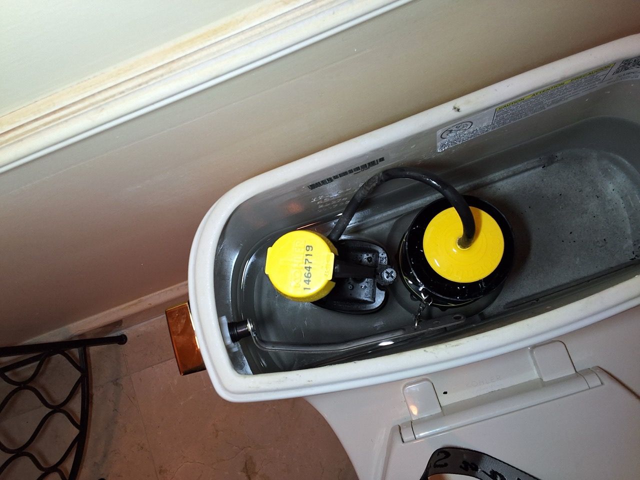 Open toilet tank with yellow float parts and water inside, beside a wall and floor cable.