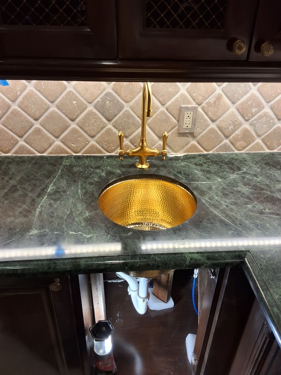 Kitchen sink with gold faucet and gold basin set into a dark green countertop, under dark cabinets