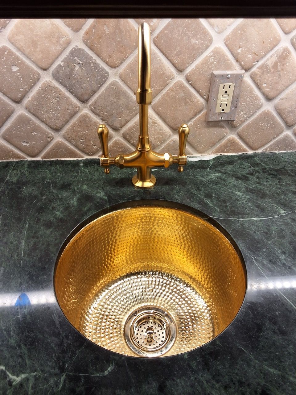 Gold kitchen sink with a tall faucet set into a dark countertop, against a tiled backsplash.