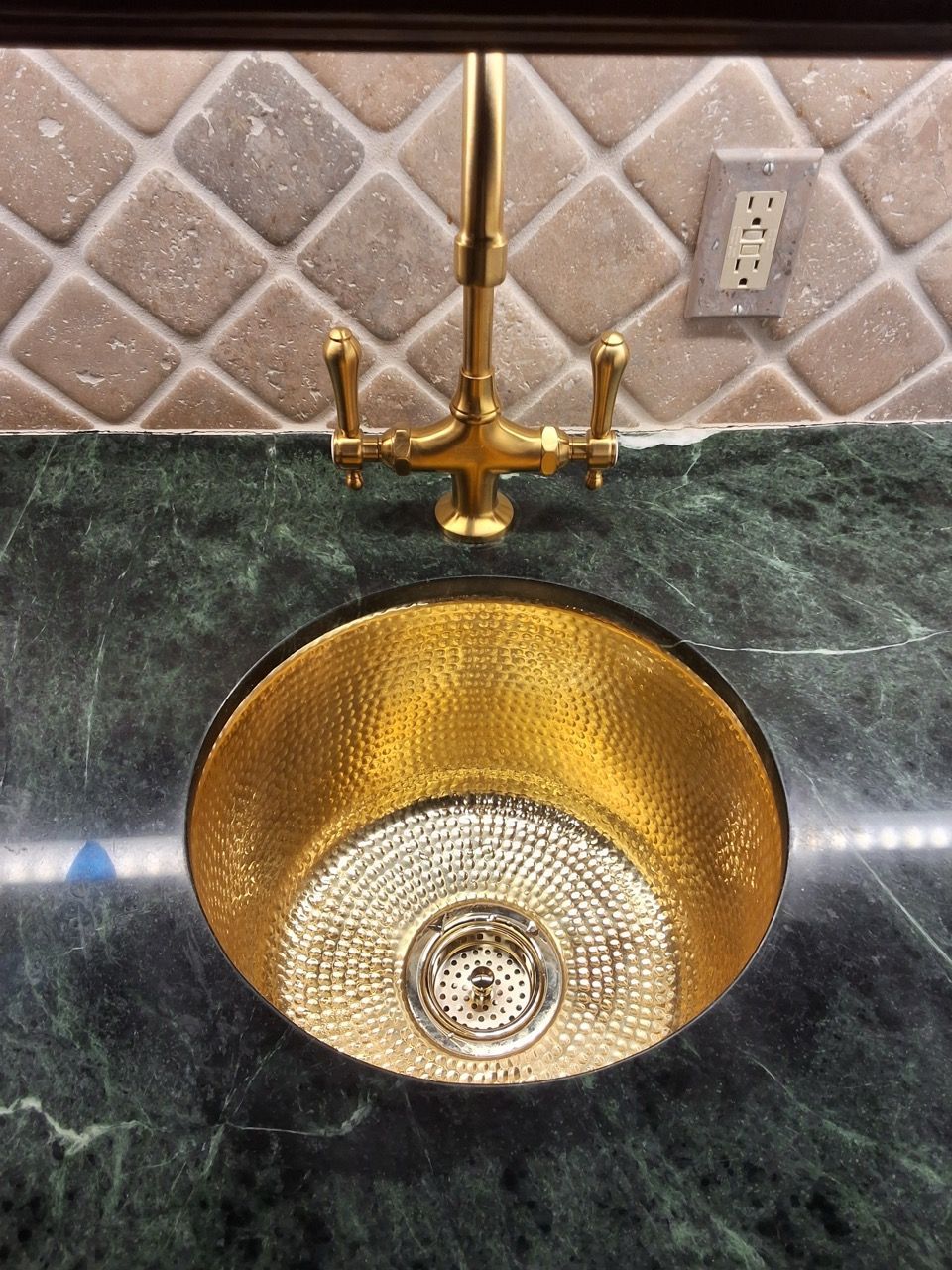 Gold vessel sink with tall faucet on a dark countertop, set against a tiled backsplash