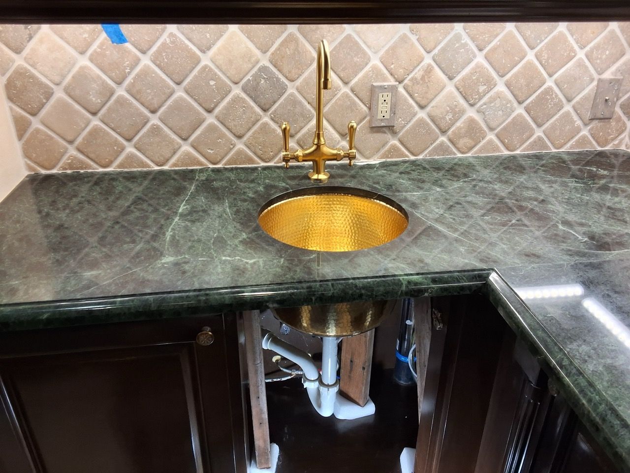 Kitchen countertop with gold sink and faucet, dark green marble surface, and beige diamond tile backsplash