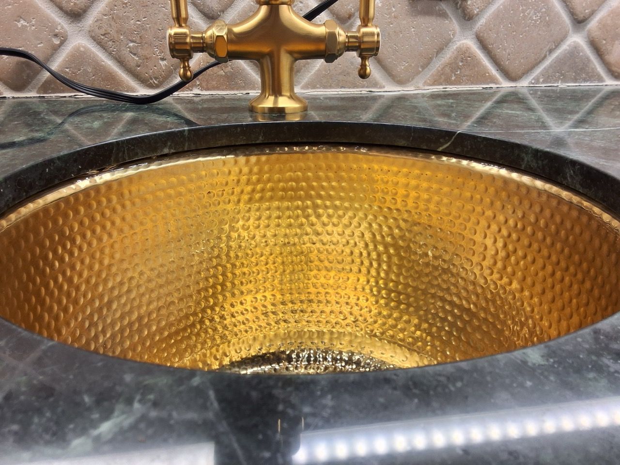 Gold hammered sink basin with black countertop and brass faucet beneath a marble backsplash