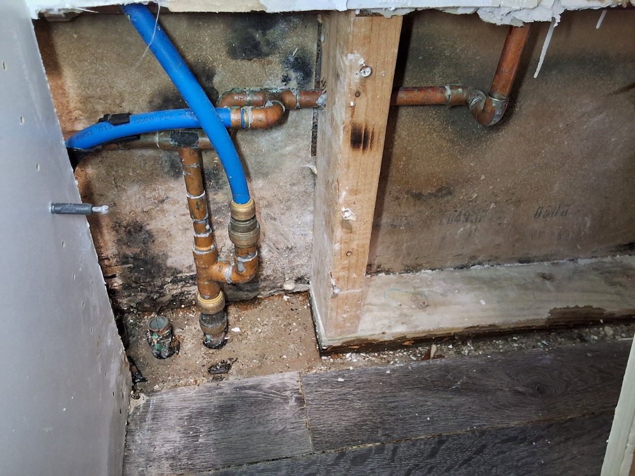 Exposed plumbing with blue pipe and copper fittings inside a wall opening, above a concrete floor