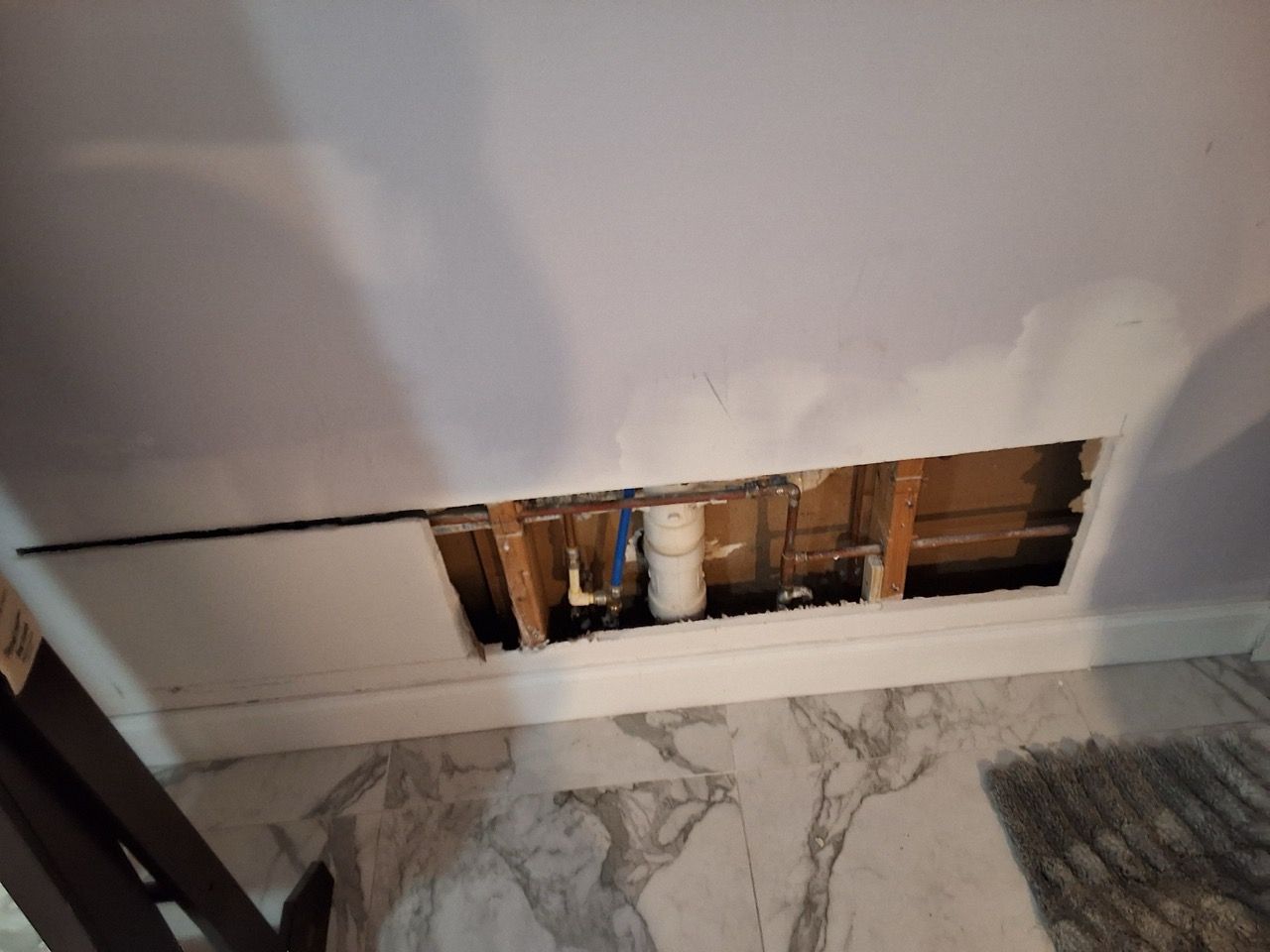 Damaged bathroom wall with a rectangular hole exposing pipes above a marble floor