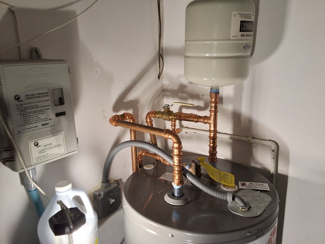 Copper pipes and water tanks in a utility room with a wall-mounted unit and a white container on the left