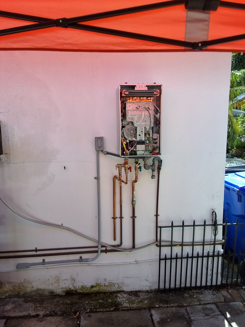 Outdoor wall-mounted utility meter with exposed pipes beneath an orange canopy, beside a fence and driveway