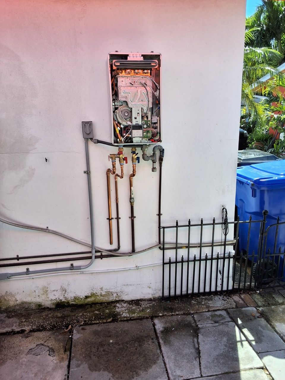 Gas meter and pipes mounted on an exterior wall beside a small fence and blue bin.
