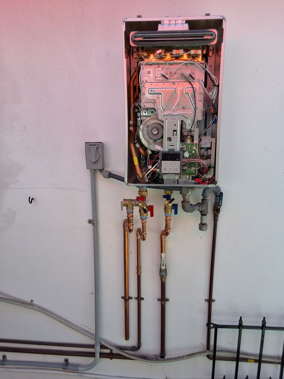 Open wall-mounted boiler with exposed copper pipes and wiring in a utility room.
