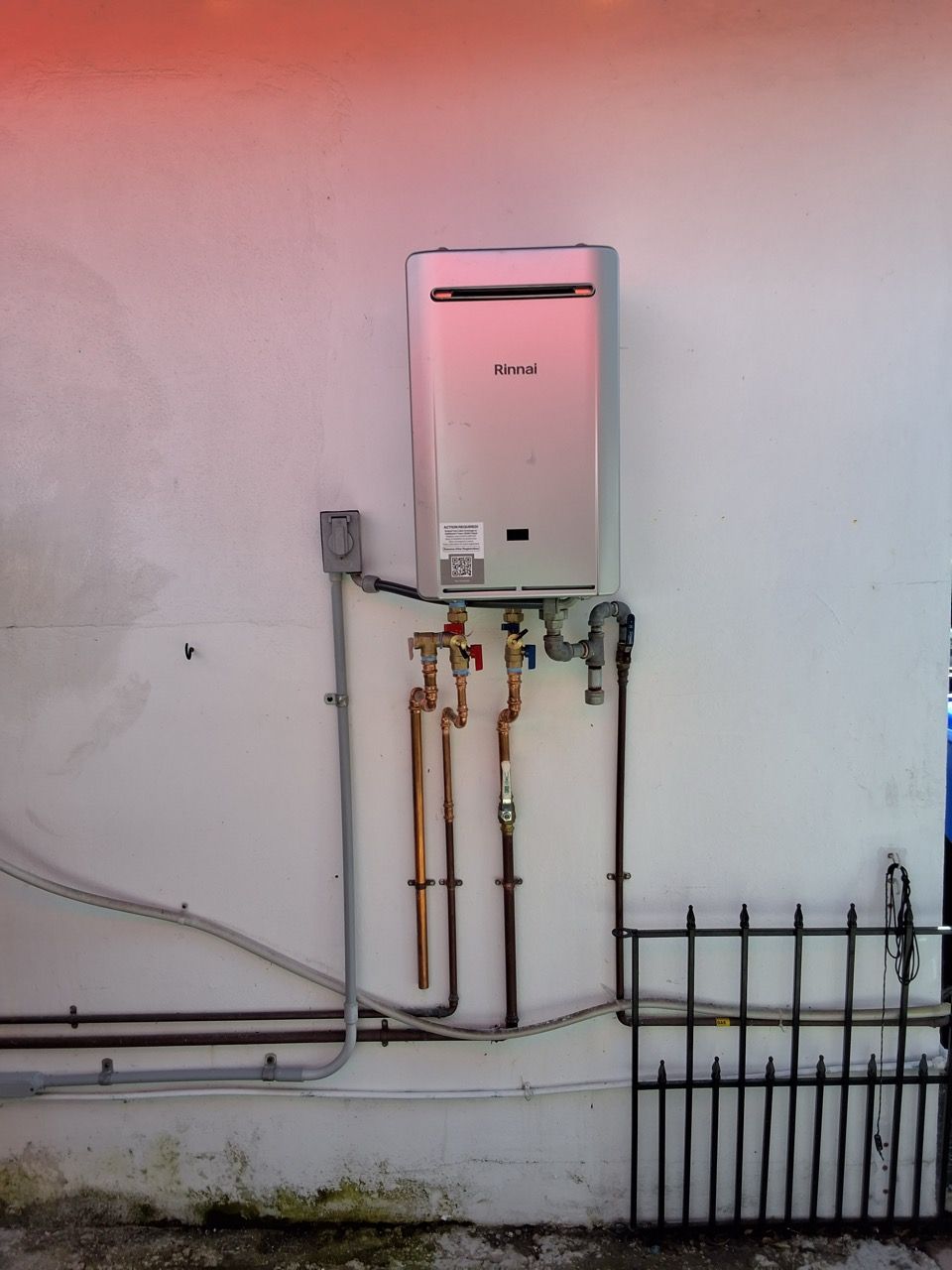 Wall-mounted utility box and pipes on an exterior wall, with a small fenced area below.