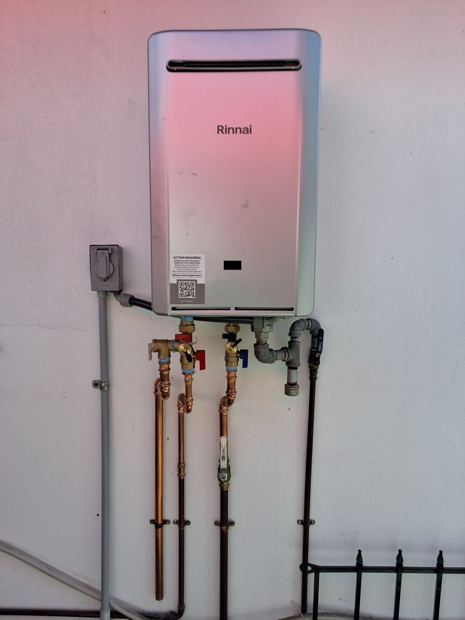Wall-mounted tankless water heater with exposed pipes and valves on a pink wall.