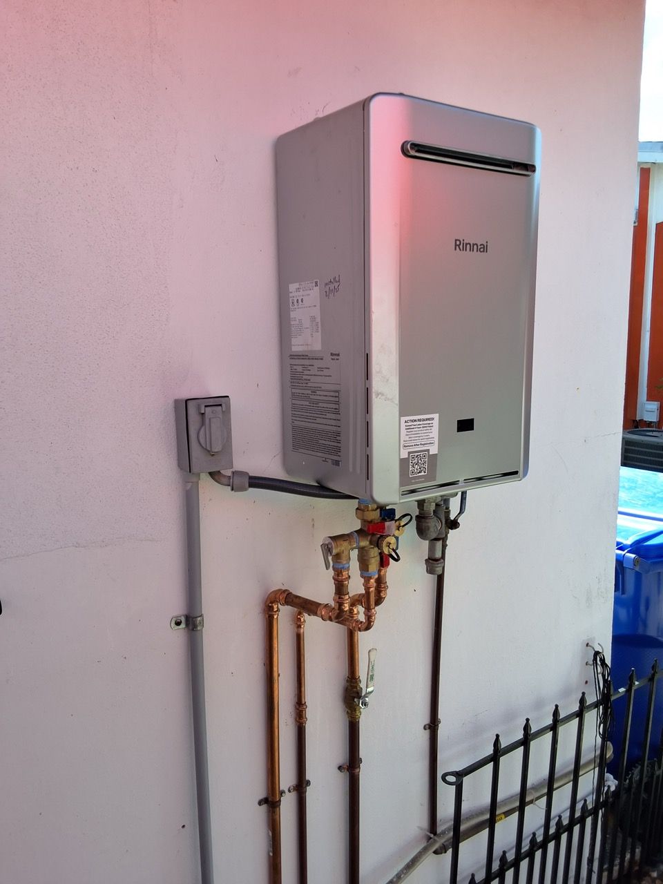 Wall-mounted outdoor boiler with exposed copper pipes beside a white stucco wall.