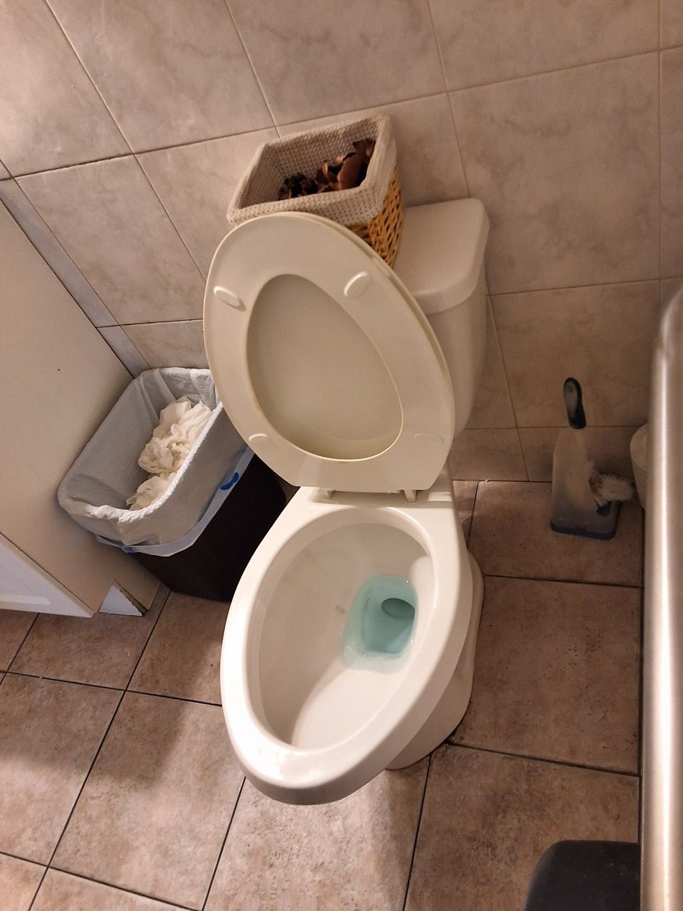 Cluttered bathroom with an open toilet, trash can, and cleaning supplies on tiled floor.