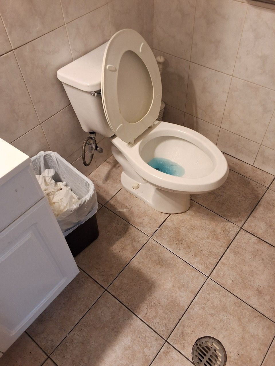 Small bathroom with an open toilet, tiled floor and walls, trash bin, and floor drain.