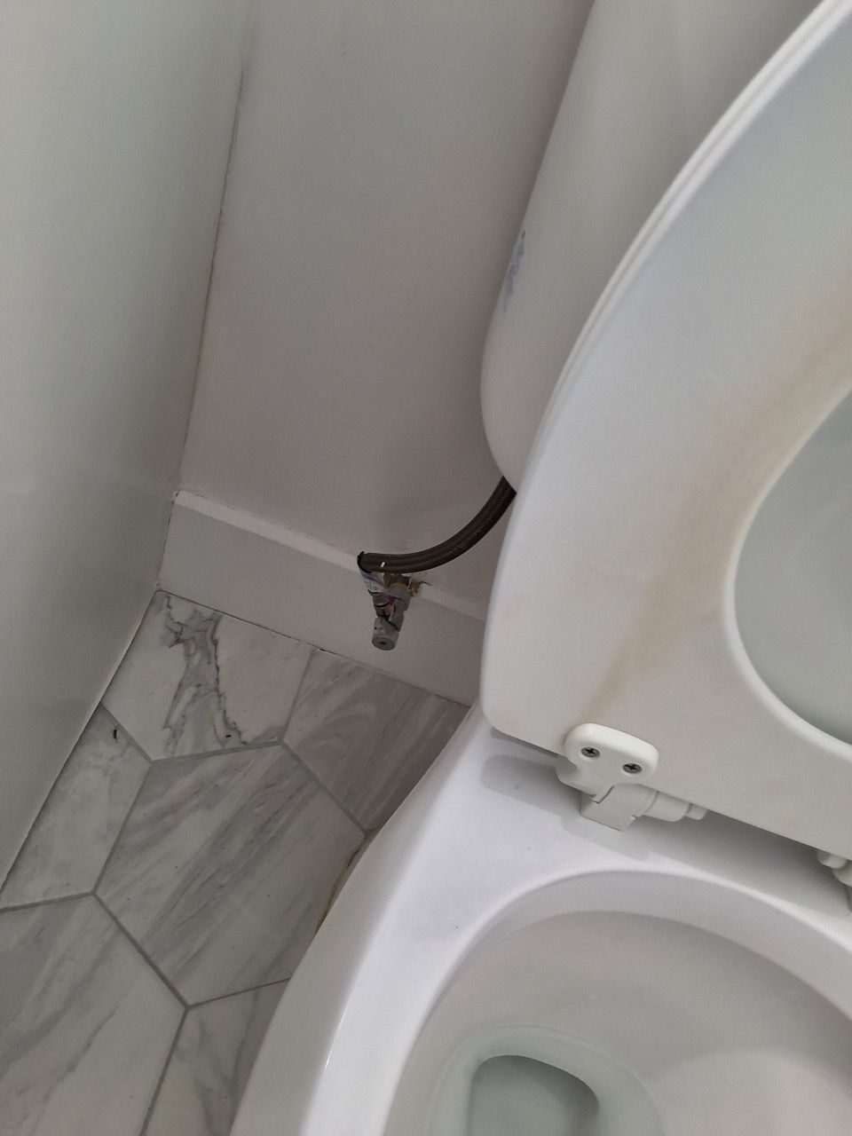 Bathroom corner with exposed hose behind a toilet, next to gray tile flooring and a white wall.