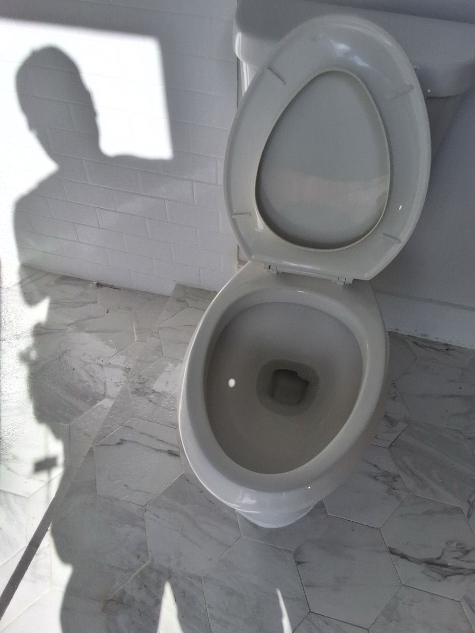 Open white toilet with lid up in a sunlit bathroom, casting a shadow on the tiled wall.
