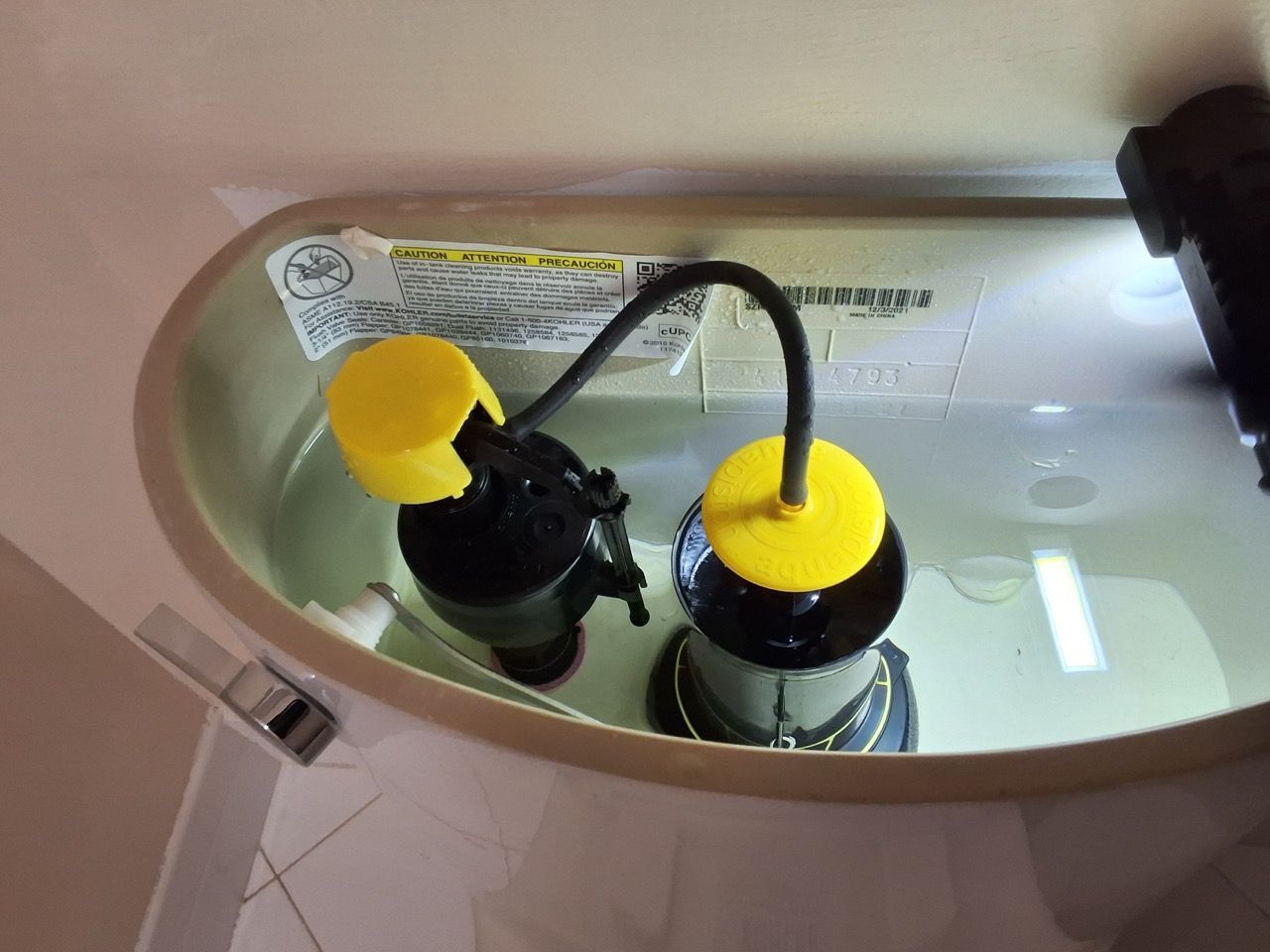 Toilet tank with two black fill valves topped by yellow caps inside a bathroom.