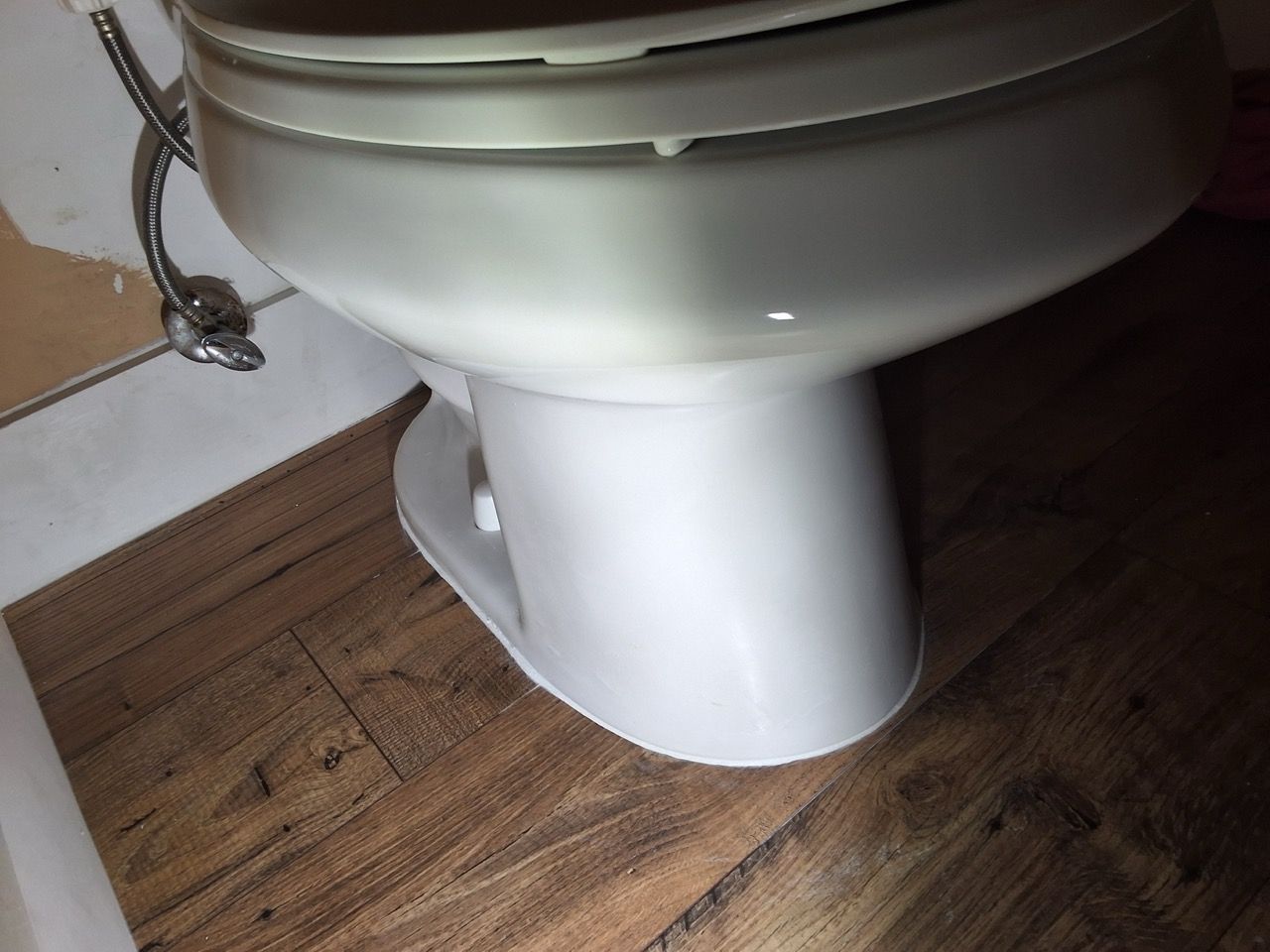 Close-up of a white toilet on wood-patterned flooring with a chrome water supply hose visible.