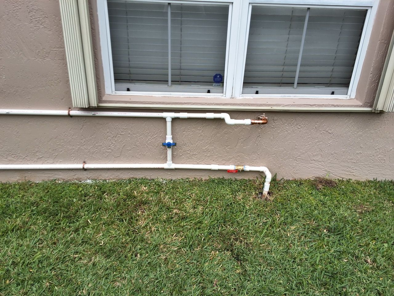 White utility pipes run along a stucco wall beneath a window above a grassy lawn.