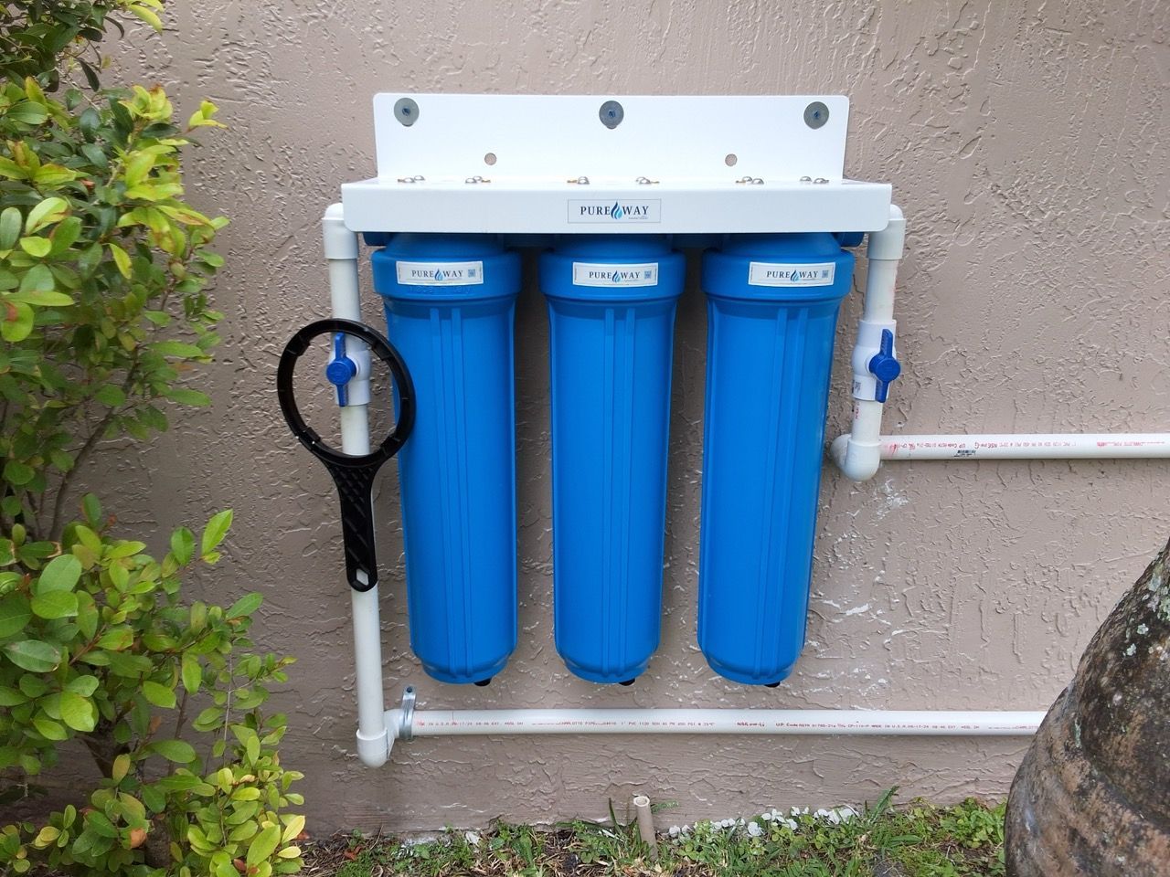 Wall-mounted water filtration system with three blue canisters and a black wrench outdoors on a stucco wall