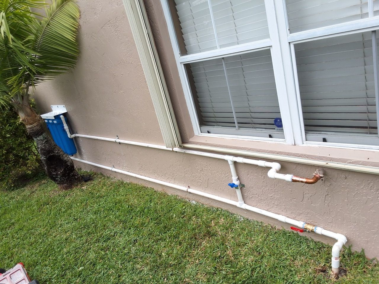 Outdoor house wall with white PVC pipes and blue utility meter beside a window and grass.
