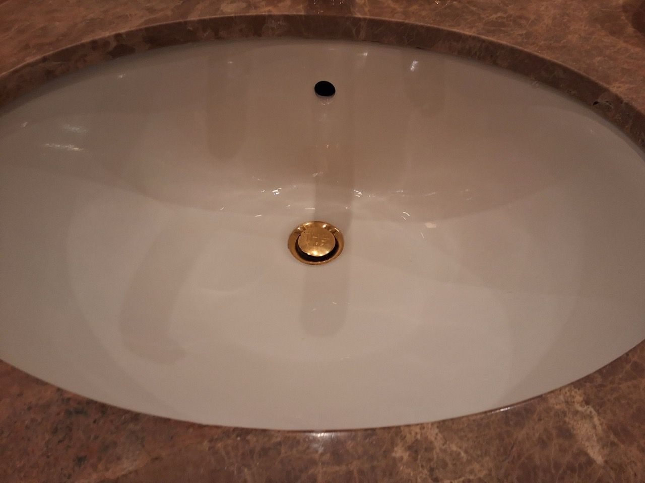 White oval sink with a bronze drain and running water in a marble countertop