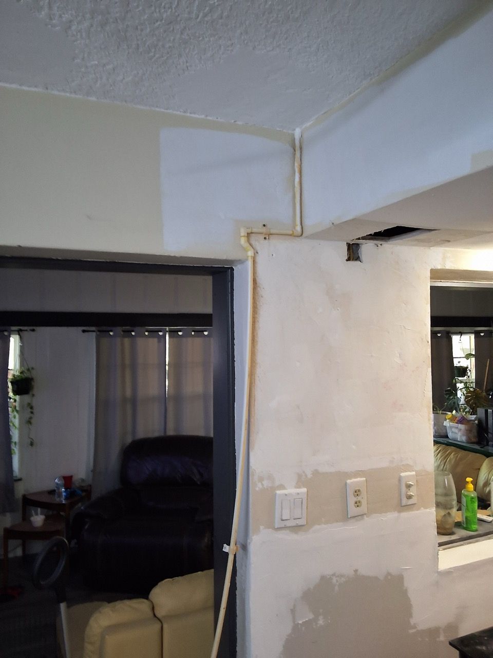 Interior wall corner with exposed drywall patch, light switch, and a pass-through opening to another room.