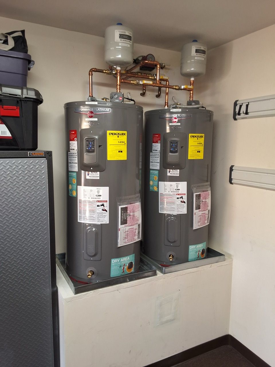 Two gray water heaters in a utility closet with pipes and labels above a white base.