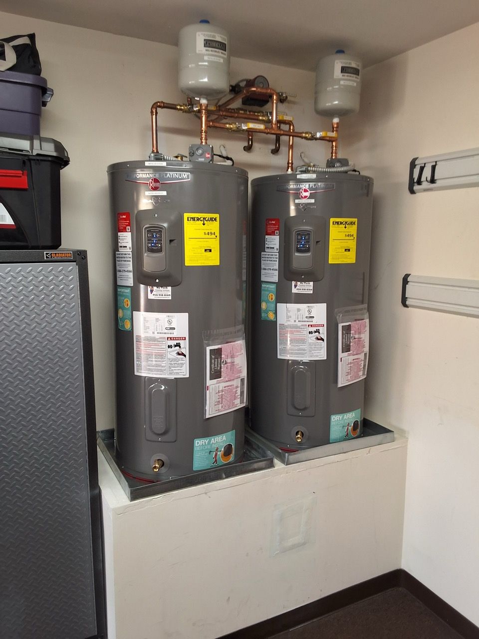 Two gray water heaters in a utility closet with pipes, valves, and a white pan beneath them