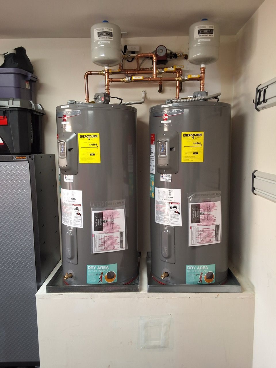 Two gray water heaters in a utility closet with pipes and expansion tanks above them