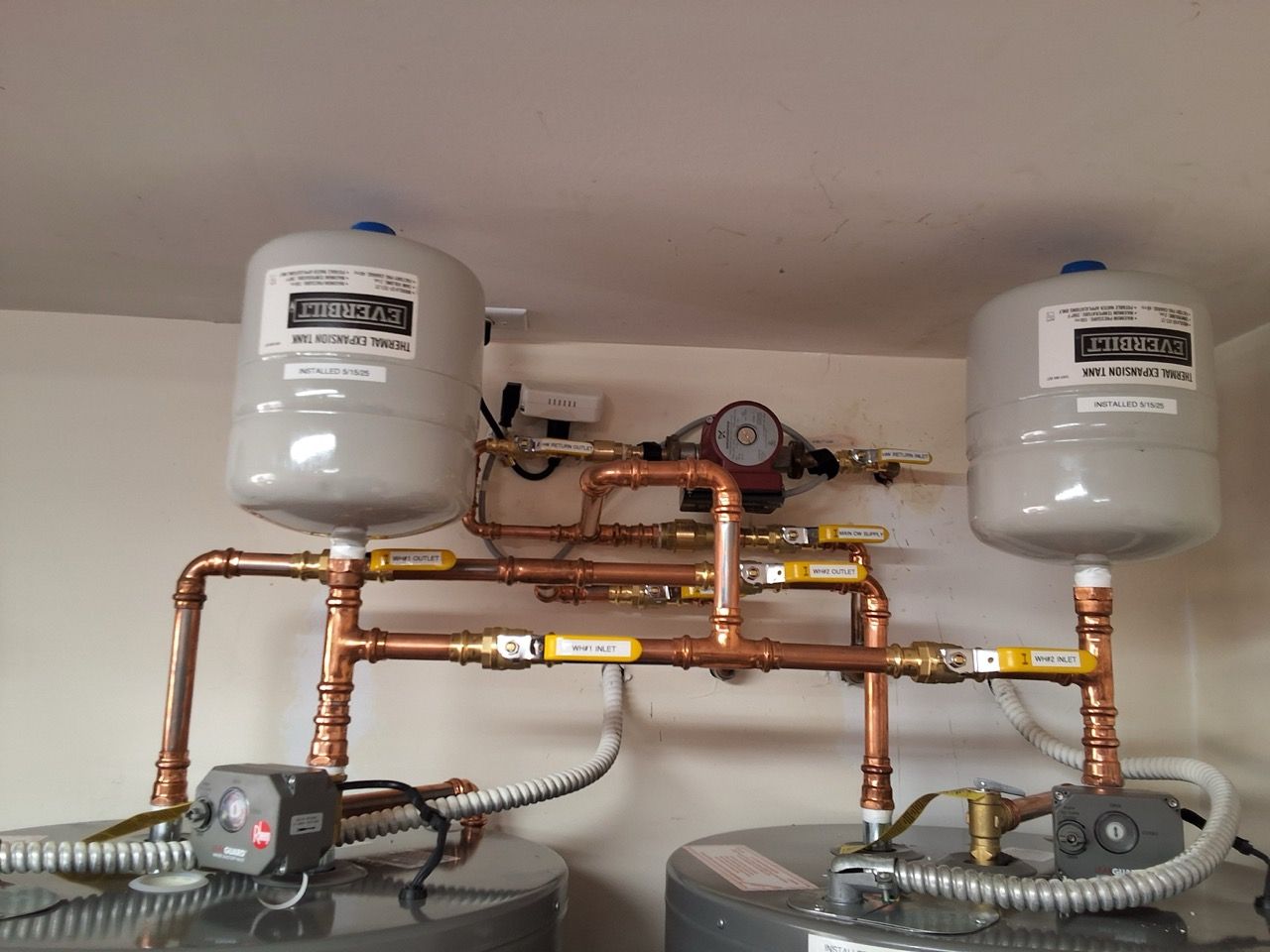 Copper pipes and two white expansion tanks mounted on a wall above heating pumps.