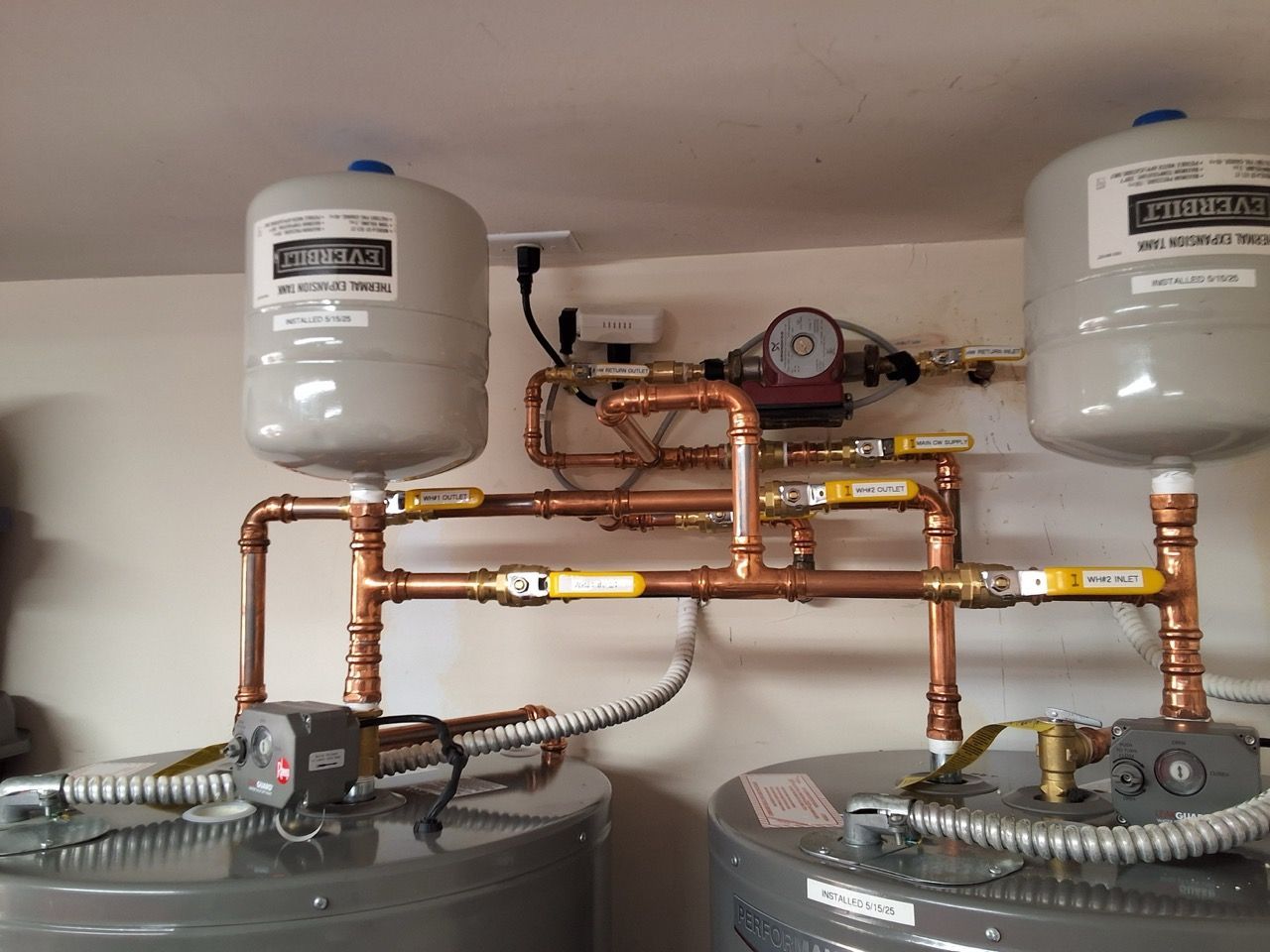 Water heater plumbing with copper pipes, two white expansion tanks, and gray tanks in a utility closet.