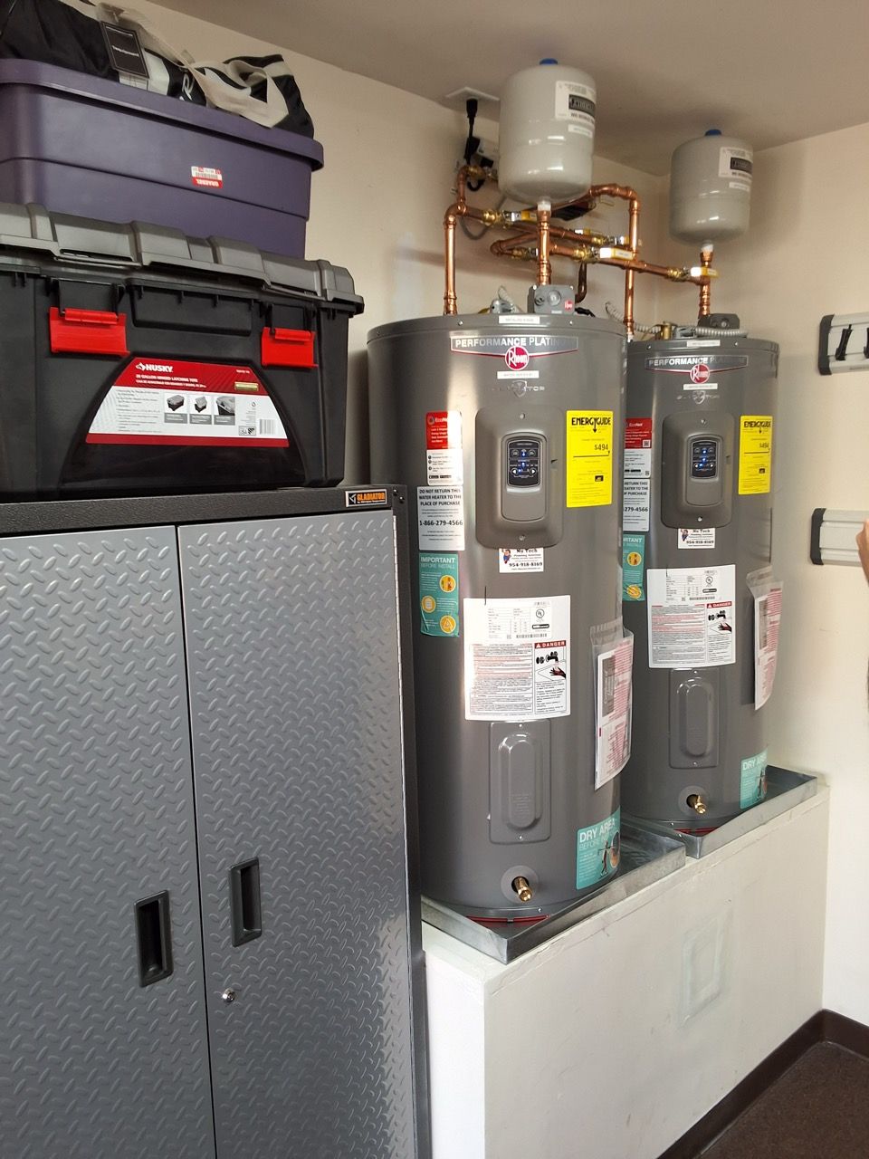 Utility room with two gray water heaters beside a tall silver cabinet and red toolbox on top