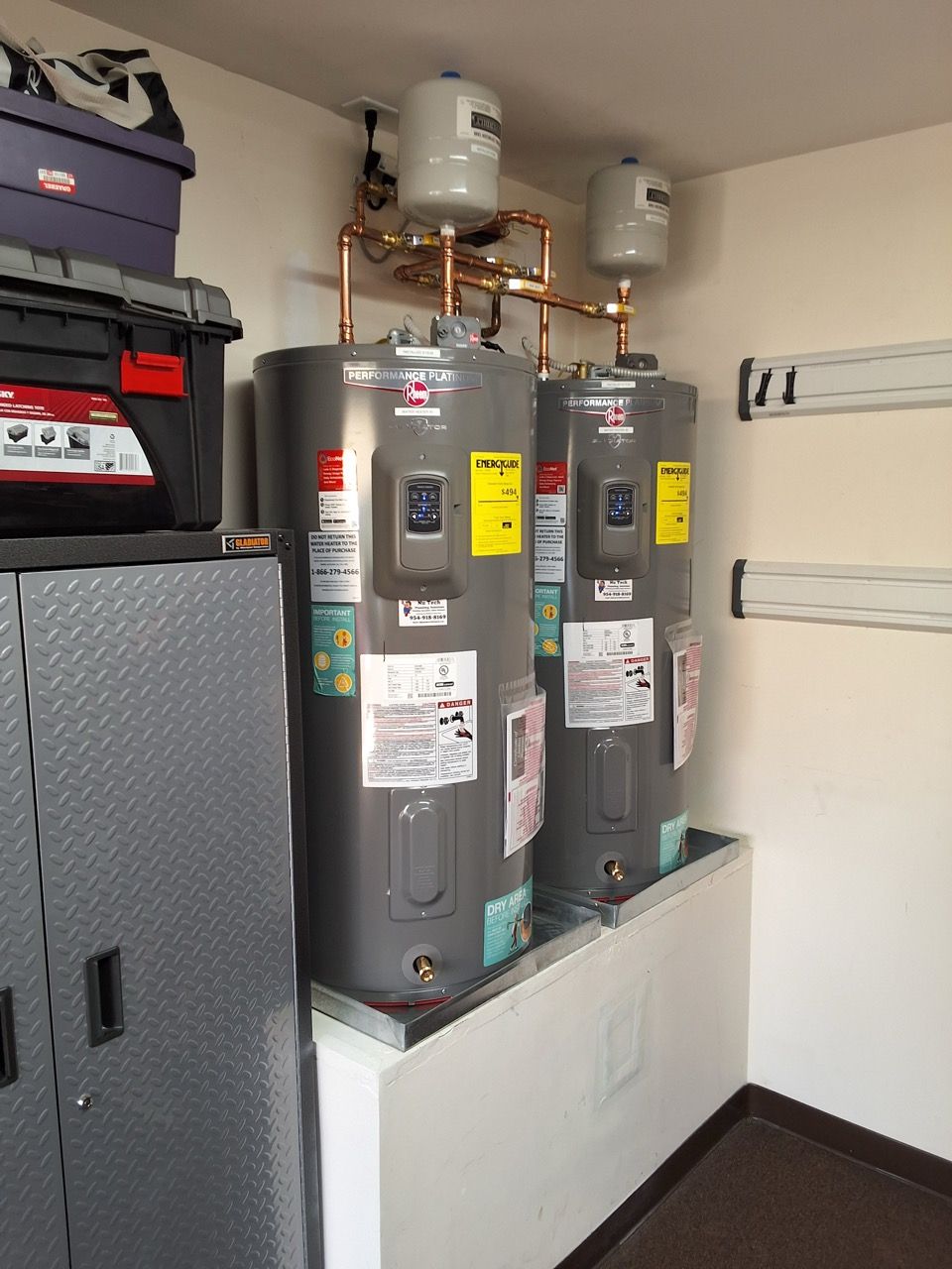 Two gray water heaters with pipes and valves in a utility room, beside a metal cabinet and wall-mounted brackets