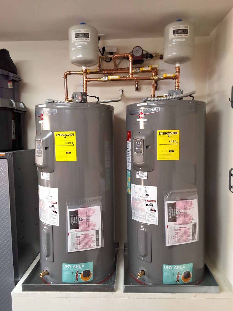 Two gray water heaters with pipes and expansion tanks in a utility closet