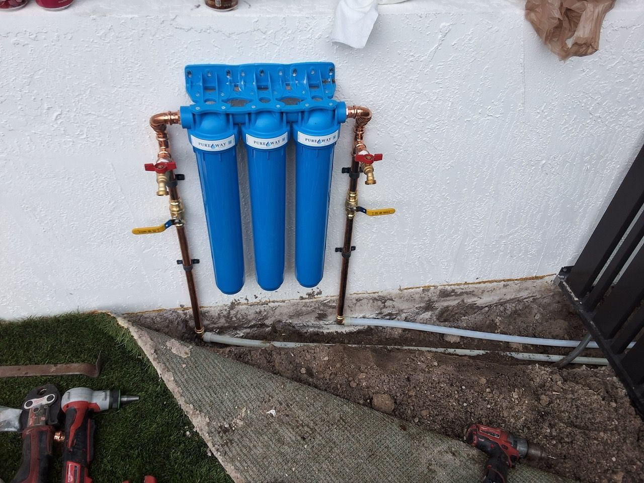 Blue water filter unit mounted on an exterior wall above a concrete trench beside grass and tools