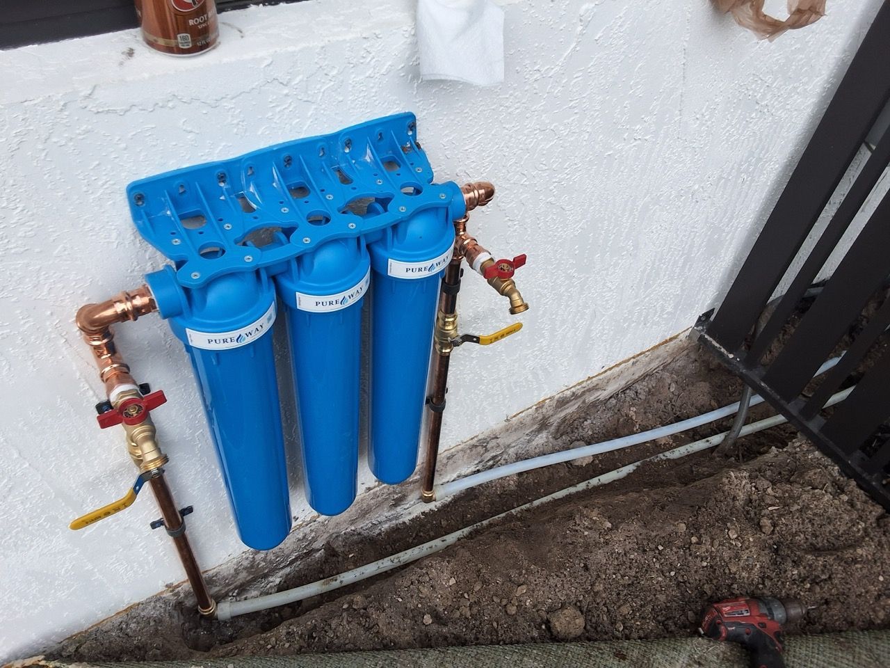 Blue water filter unit with four cartridges mounted on an exterior wall, with copper pipes and valves.