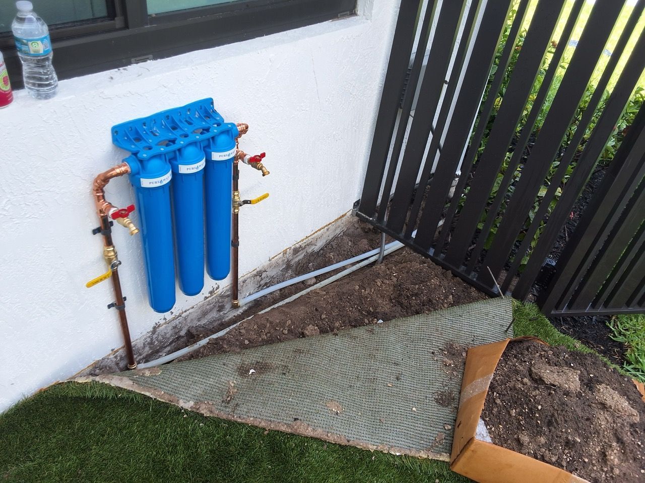 Blue water filter pipes beside a house wall, with gravel and a small lawn edge in the corner.
