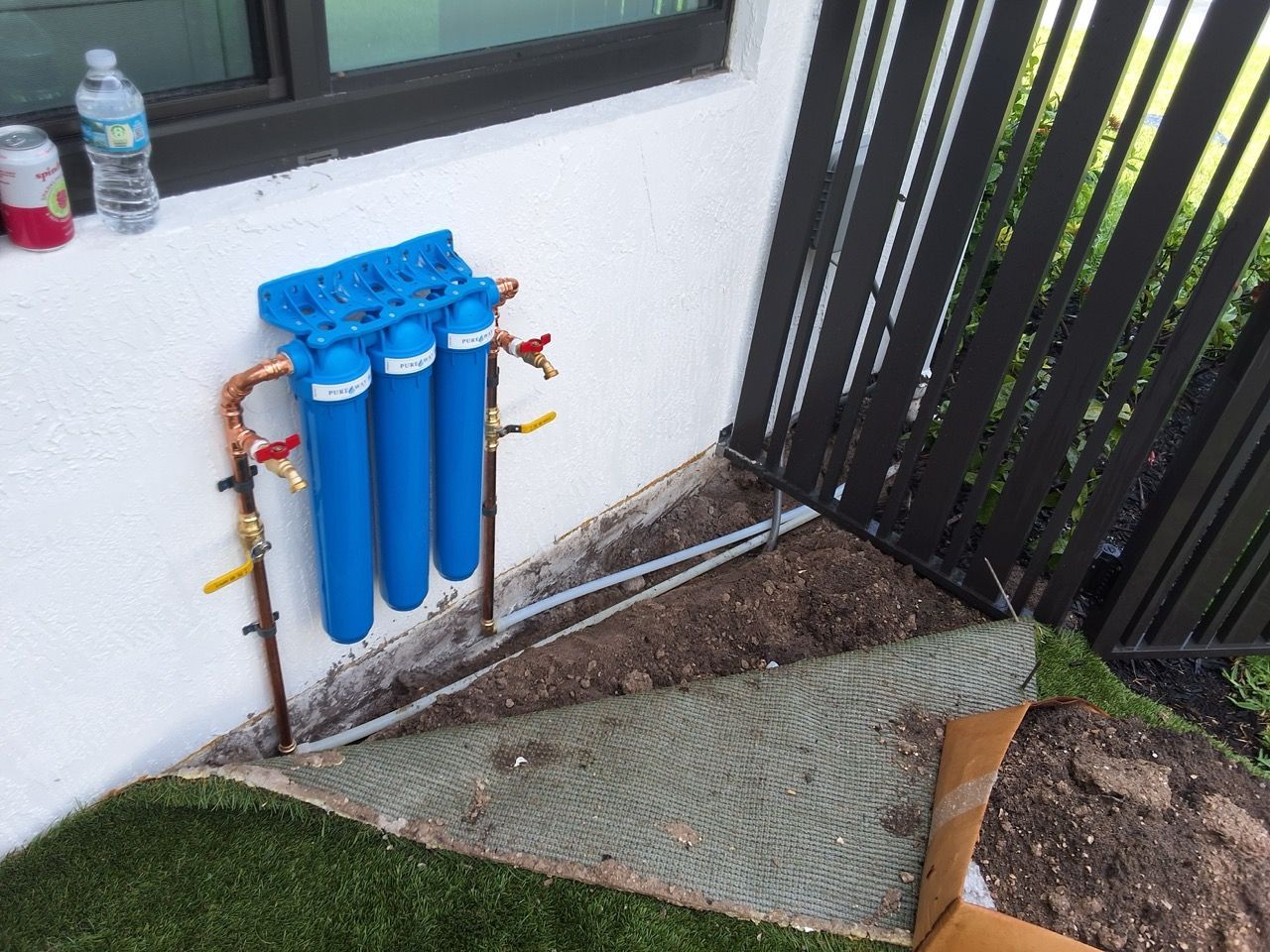Blue water filter cylinders mounted on an exterior wall beside a window and fence, over a gravel area