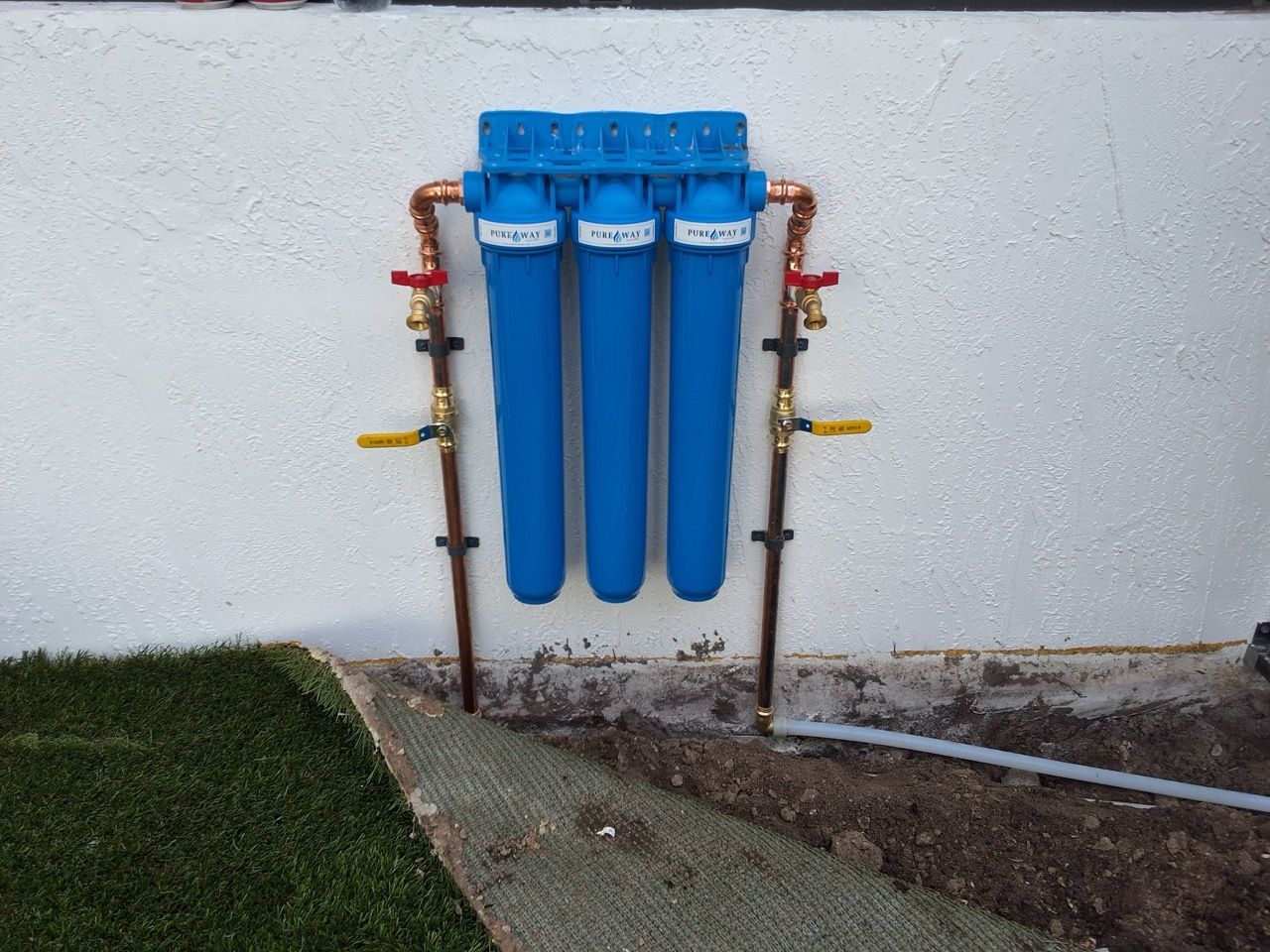 Three blue water filter cartridges mounted on an exterior wall with pipes and valves