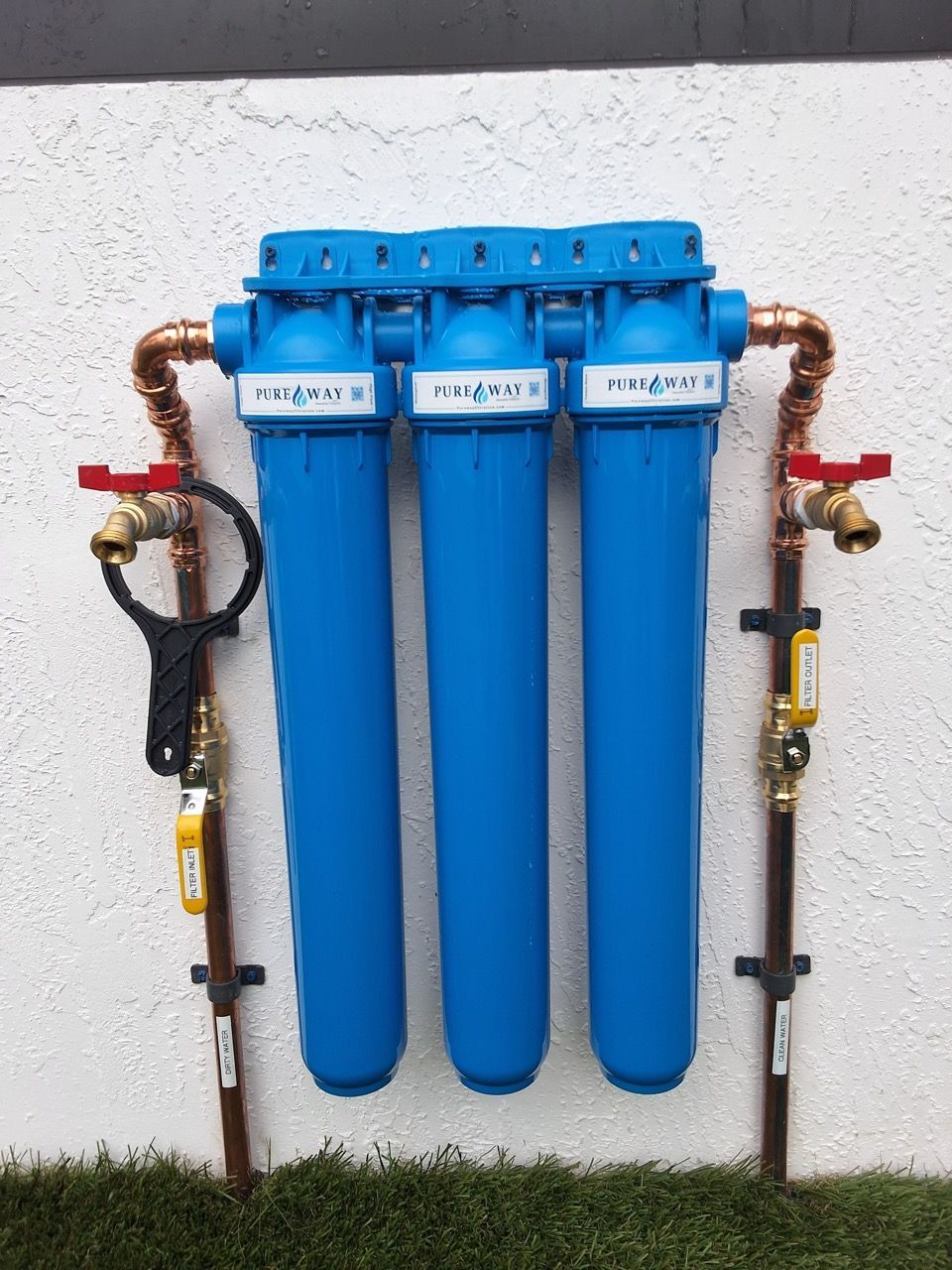 Blue water filtration system with three canisters, pipes, and red valves mounted on a white wall.