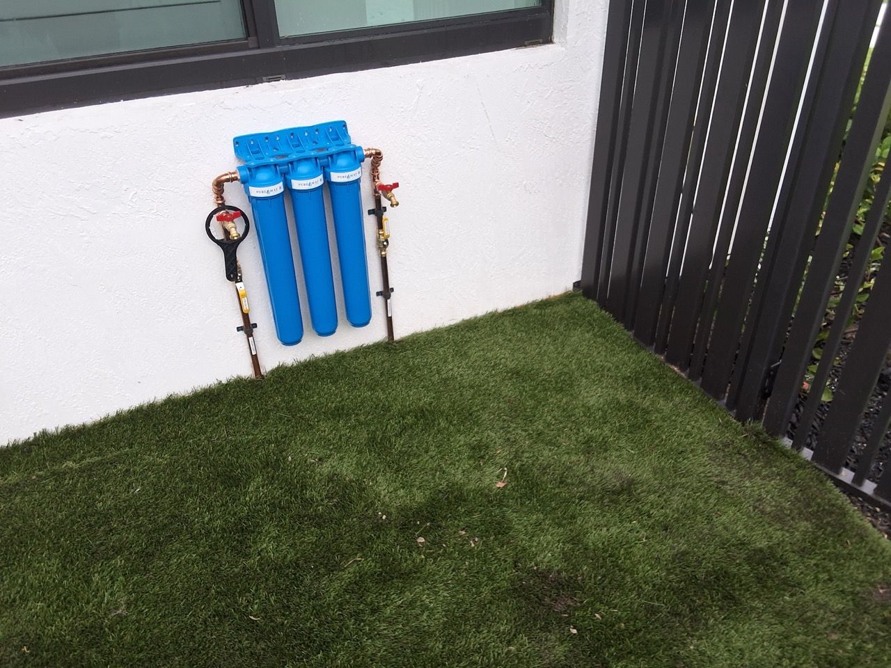 Blue water filters mounted on an outdoor wall beside a grassy yard and black fence