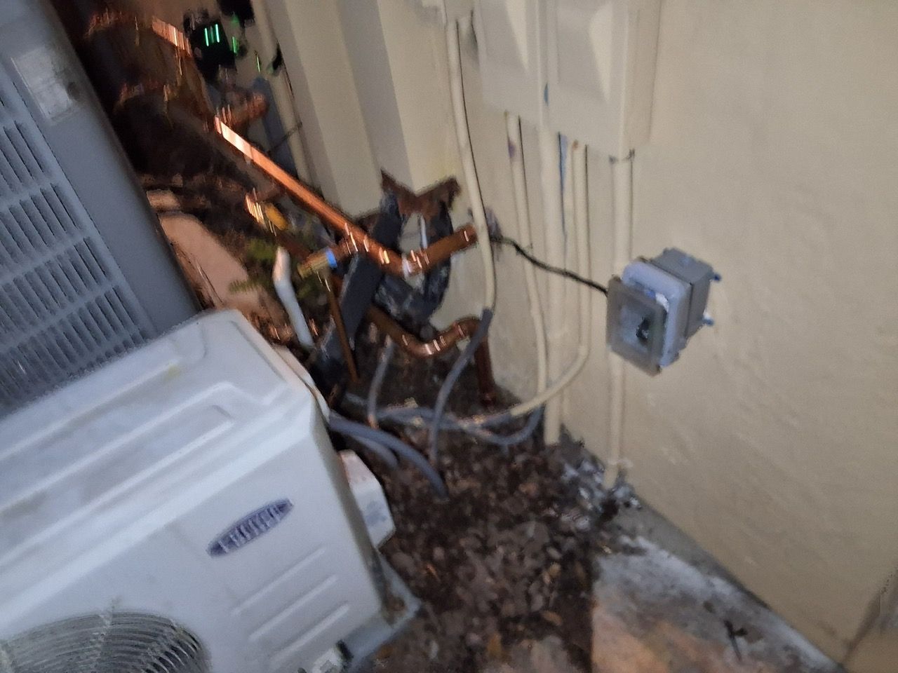 Damaged outdoor HVAC unit with exposed copper pipes and debris beside a beige wall outlet