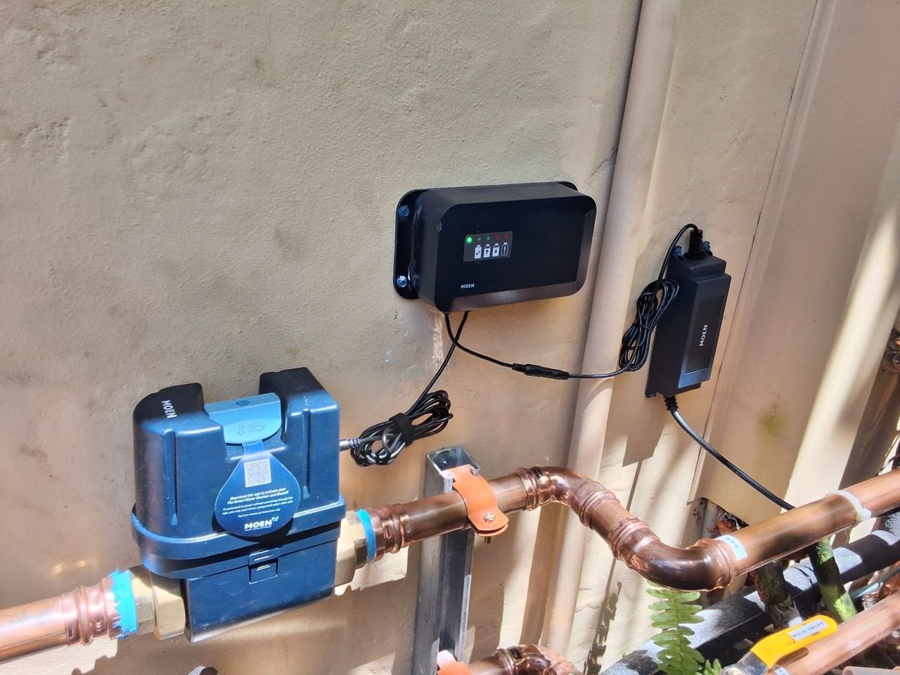 Blue water meter and pipes mounted on an exterior wall, with a black control box nearby.